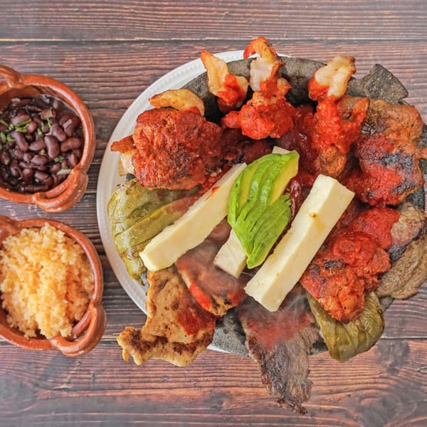 Steaming molcajete filled with grilled meats, shrimp, cheese, avocado, and peppers, with sides of rice and beans.