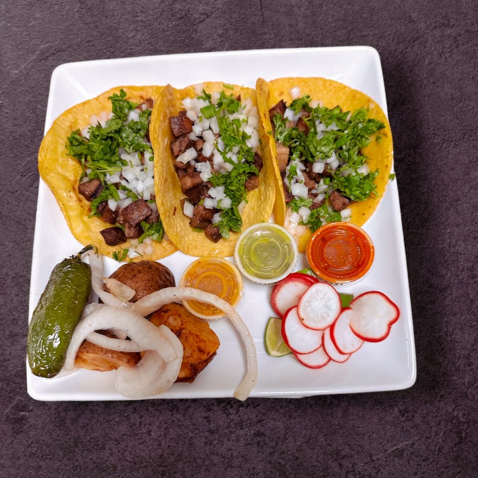 Three tacos with meat, cilantro, and onions, served with sauces, grilled pepper, and radishes.
