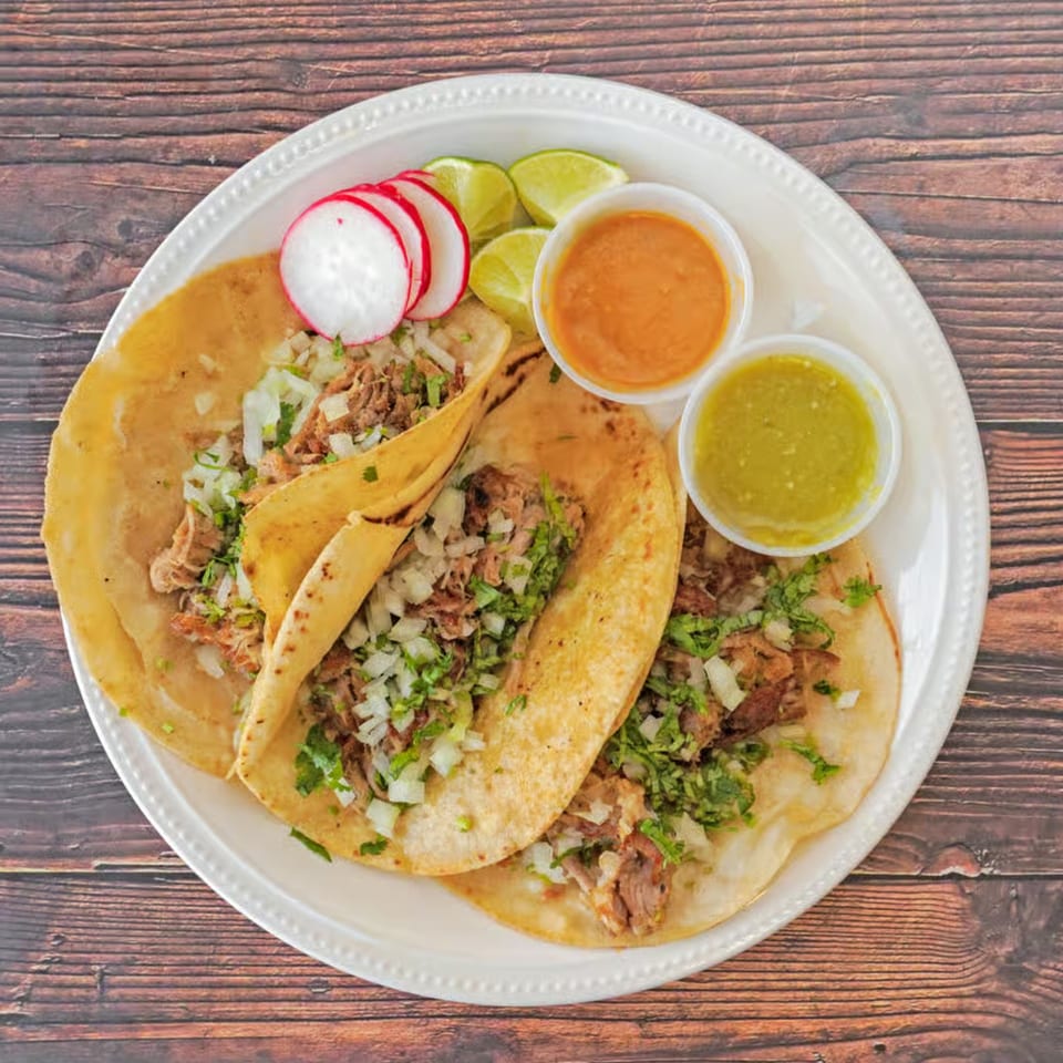 Three street tacos with meat, onions, and cilantro, served with radishes, limes, and two sauces.