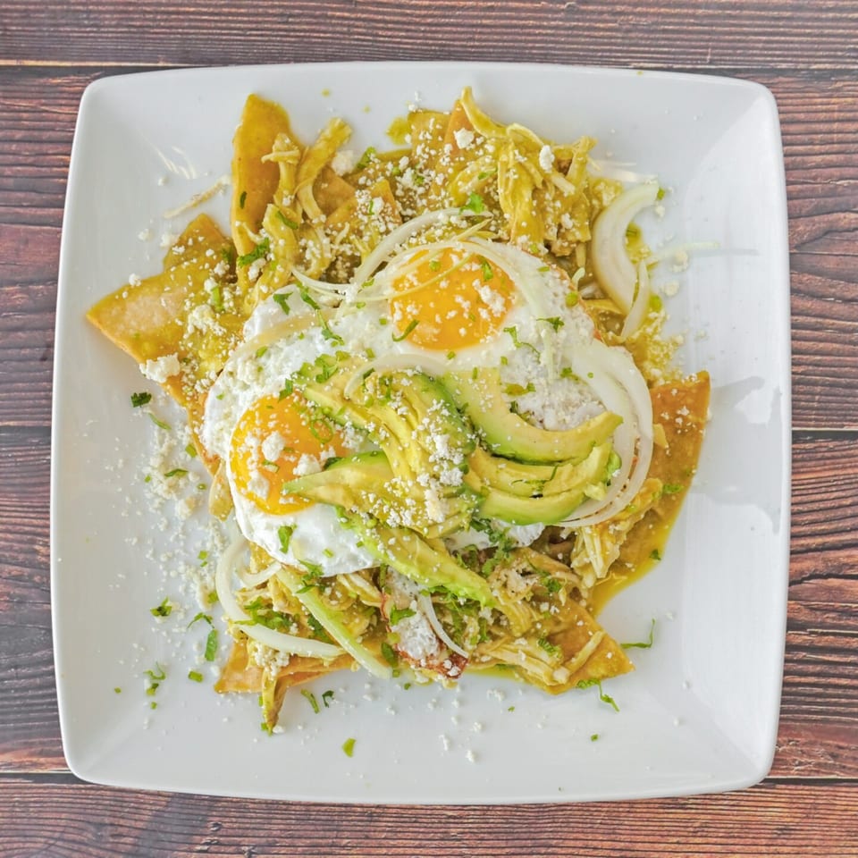 Chilaquiles with green sauce, shredded chicken, two fried eggs, avocado, onions, and cheese.